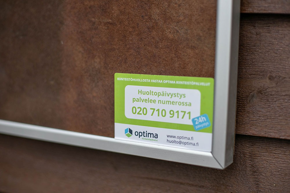 Ilmoitustaululla Optima Kiinteistöpalveluiden tarra, jossa näkyy huoltopäivystyksen yhteystiedot. Kiinteistöhuolto ja kiinteistöpalvelu Helsinki Vantaa Uusimaa